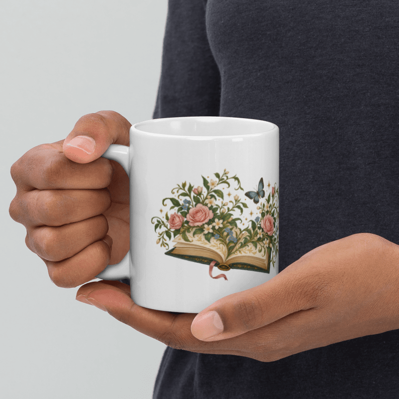 hands holding a white ceramic Where Stories Bloom mug with the floral book illustration facing out